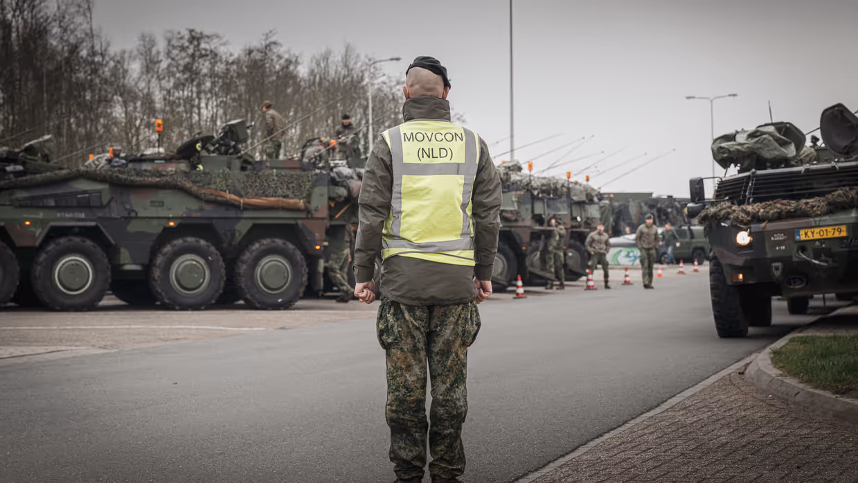 Militair met geel hesje van Movcon voor geparkeerde militaire voertuigen.