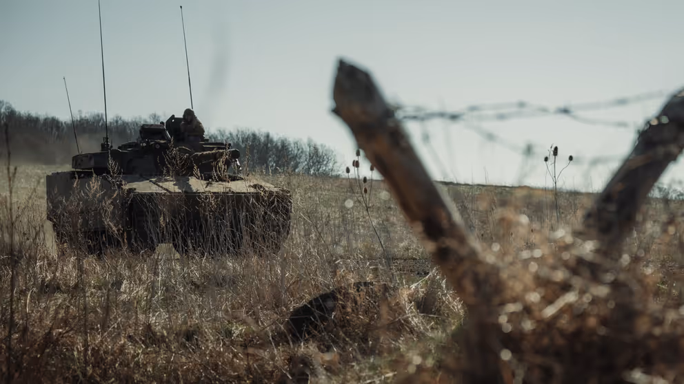 Een tank op een veld. Op de voorgrond 2 schuine palen in V-vorm met prikkeldraad.