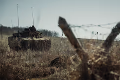 Een tank op een veld. Op de voorgrond 2 schuine palen in V-vorm met prikkeldraad.