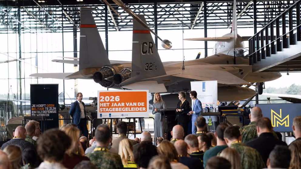 Publiek kijkt naar podium met mensen en een oranje bord. Daarachter hangt een gevechtsvliegtuig.