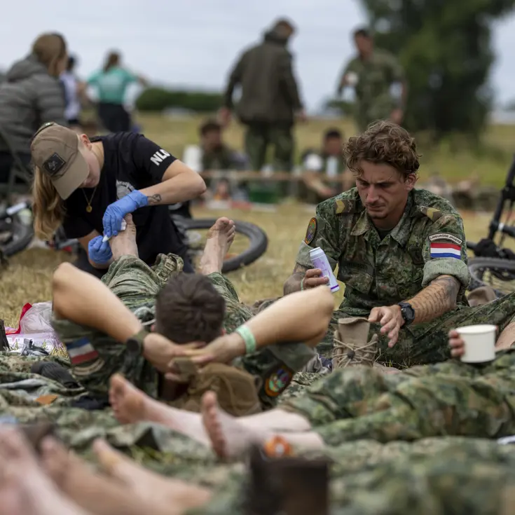 Een vrouw behandelt de voeten van een militair, die in het gras op zijn rug tussen collega’s ligt.  