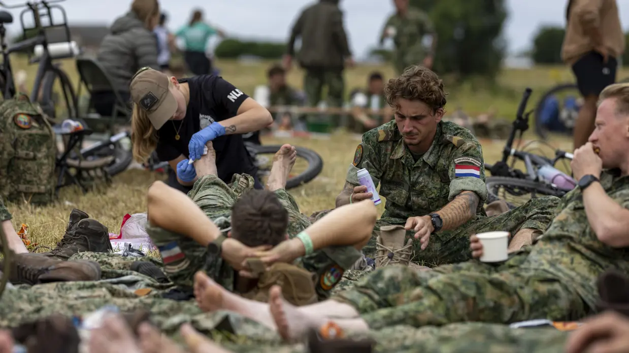 Een vrouw behandelt de voeten van een militair, die in het gras op zijn rug tussen collega’s ligt.  