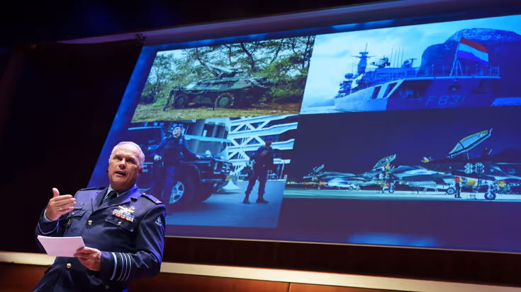 Eichelsheim staat op het podium voor een groot scherm en spreekt de zaal toe. 