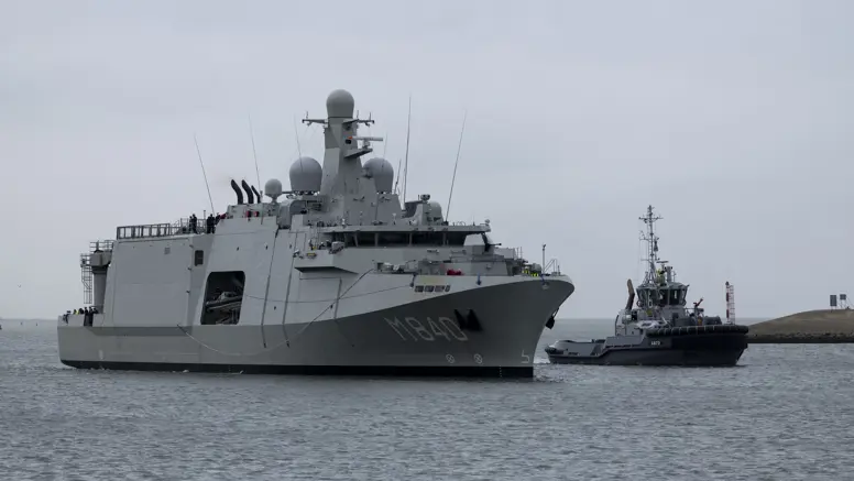 Een grijs marineschip vaart op een kalme zee, daarnaast een sleepboot. 