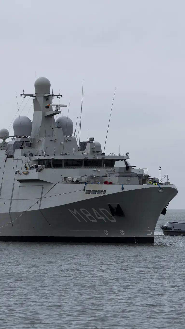 Een grijs marineschip vaart op een kalme zee, daarnaast een sleepboot. 