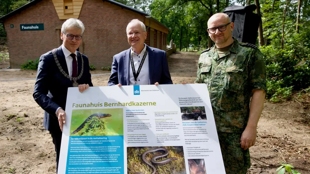 3 mannen voor een Faunahuis. In hun handen een groot bord met informatie over het Faunahuis op de Bernhardkazerne