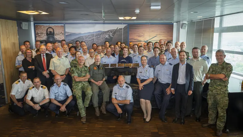 Groepsfoto van militairen en burgers rondom een schaalmodel van een satelliet.