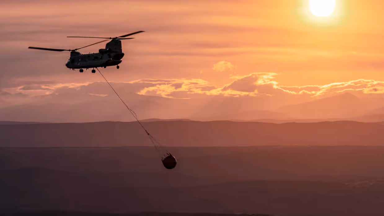 Vliegende Chinook-helikopter met bambi bucket eraan hangend. De lucht is zalmkkleurig en de zon is zichtbaar.