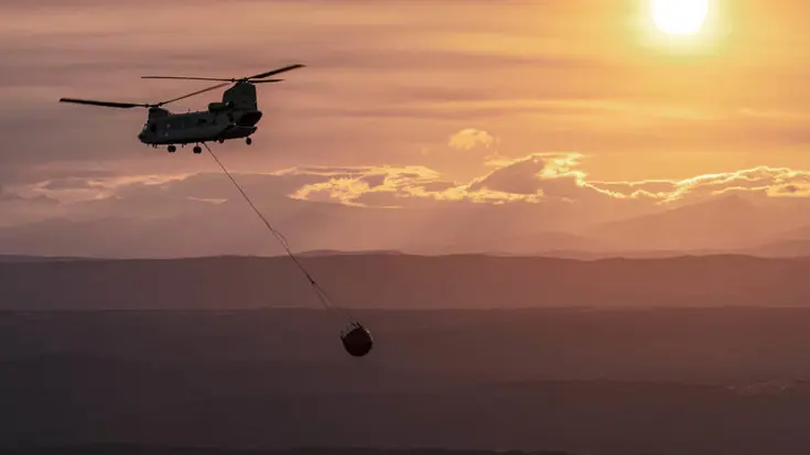 Vliegende Chinook-helikopter met bambi bucket eraan hangend. De lucht is zalmkkleurig en de zon is zichtbaar.