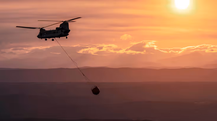 Vliegende Chinook-helikopter met bambi bucket eraan hangend. De lucht is zalmkkleurig en de zon is zichtbaar.