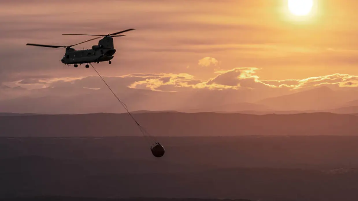 Vliegende Chinook-helikopter met bambi bucket eraan hangend. De lucht is zalmkkleurig en de zon is zichtbaar.
