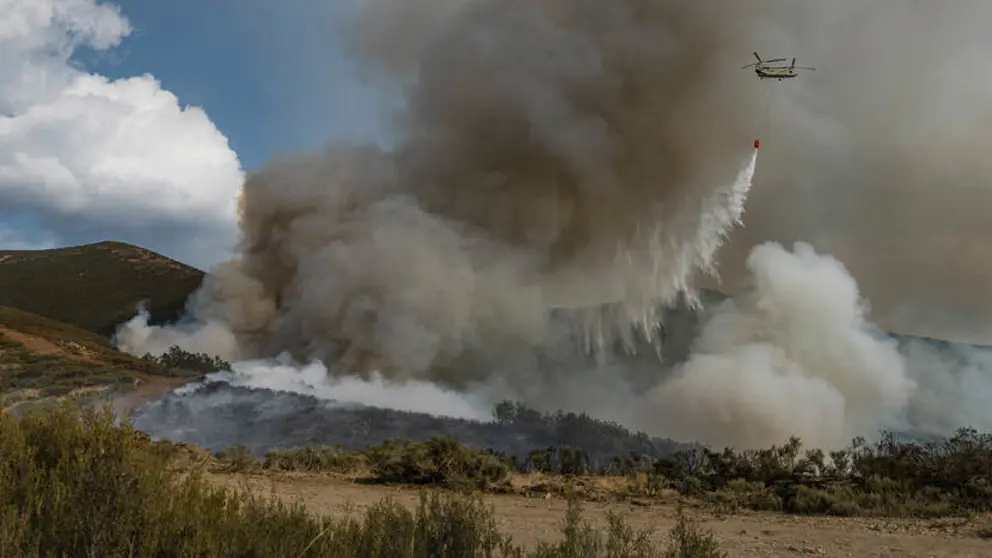 Chinook-helikopter die water uit een oranje bambi bucket laat vallen op brandend terrein.