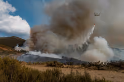Chinook-helikopter die water uit een oranje bambi bucket laat vallen op brandend terrein.