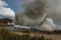 Chinook-helikopter die water uit een oranje bambi bucket laat vallen op brandend terrein.