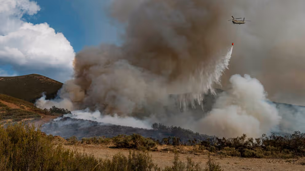 Chinook-helikopter die water uit een oranje bambi bucket laat vallen op brandend terrein.
