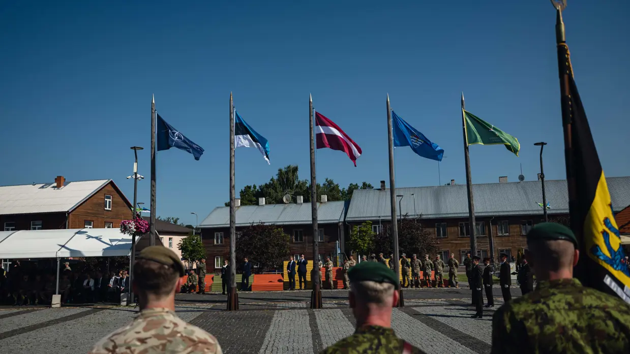 Drie militairen op de rus gezien kijkend naar wapperende landenvlaggen bij de overdracht.