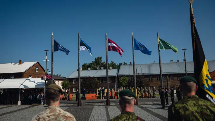 Drie militairen op de rus gezien kijkend naar wapperende landenvlaggen bij de overdracht.