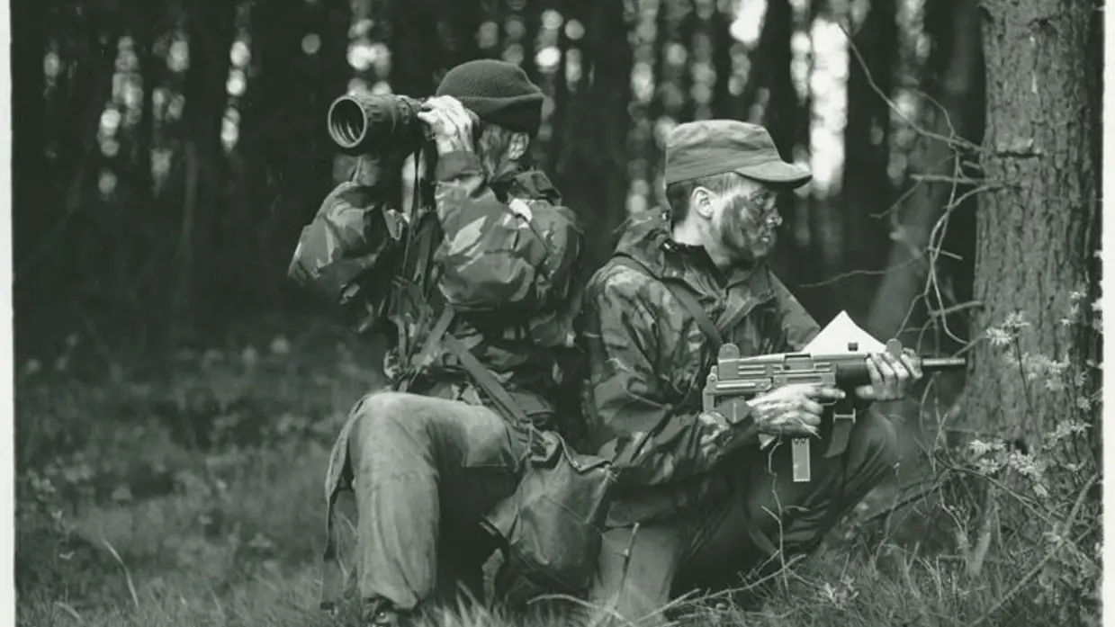 Militairen in bos. 1 met wapen en 1 met verrekijker.