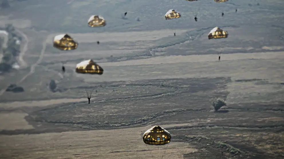 Meerdere parachutisten dalen met ronde parachutes af boven een uitgestrekt, landelijk landschap.