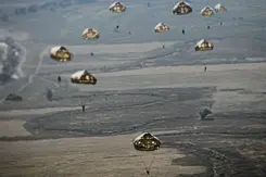 Meerdere parachutisten dalen met ronde parachutes af boven een uitgestrekt, landelijk landschap.