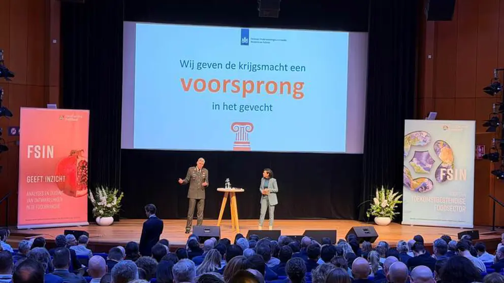 Luitenant-generaal Jan-Willem Maas staat op een podium met achter hem een groot scherm. Op de voorgrond van de foto achterhoofden van mensen in de zaal.