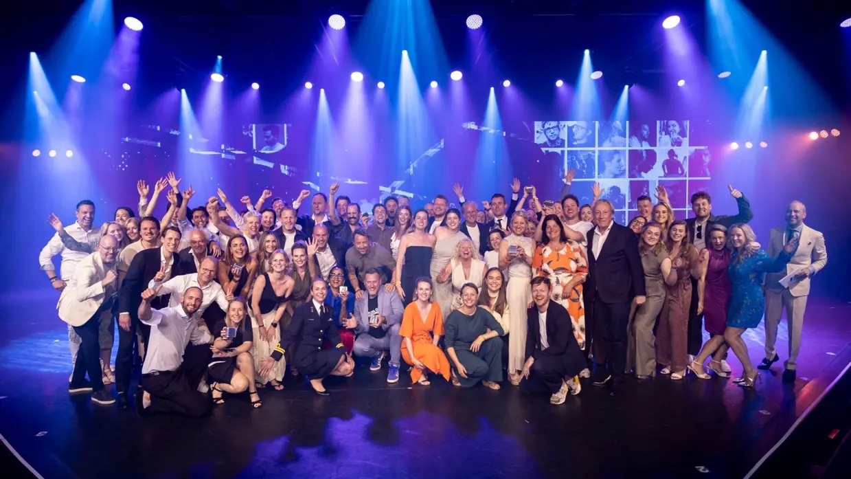 Een grote groep feestelijk geklede enthousiaste mensen op een blauw verlicht podium poseert voor de camera. Te midden van hen één vrouwelijke marineofficier.