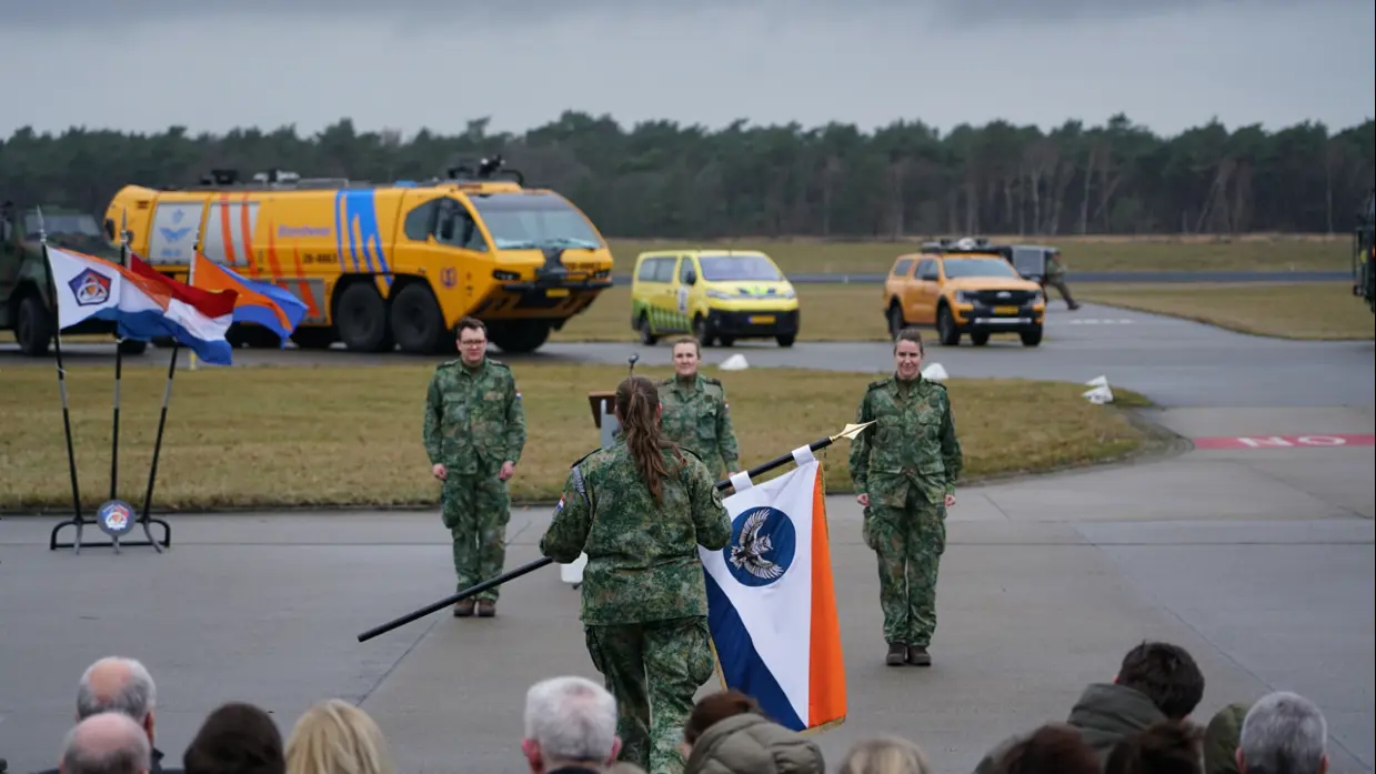 Vrouwelijke militair met vlag in de hand tegenover drie militairen op een platform. Achter hen militaire voertuigen waaronder een bluswagen. Op de voorgrond de achterhoofden van publiek.