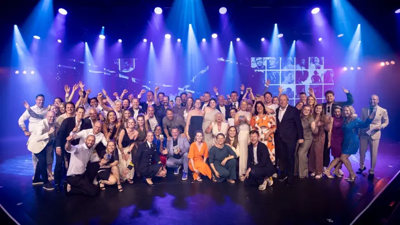 Een grote groep feestelijk geklede enthousiaste mensen op een blauw verlicht podium poseert voor de camera. Te midden van hen één vrouwelijke marineofficier.