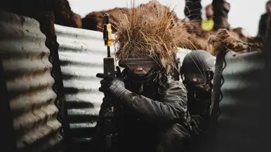 Links: Luchtfoto van loopgraven op militair oefenterrein. Rechts: Militair met wapen hurkt in loopgraaf waarvan de wanden met golfplaten zijn bekleed. Op zijn helm een bos stro als camouflage.