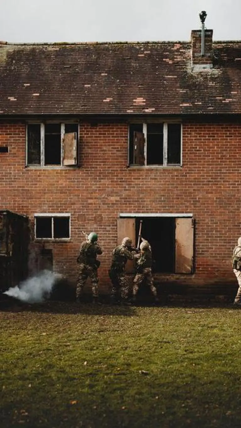 4 soldaten in camouflage benaderen een verlaten bakstenen gebouw, rook komt uit een raam.