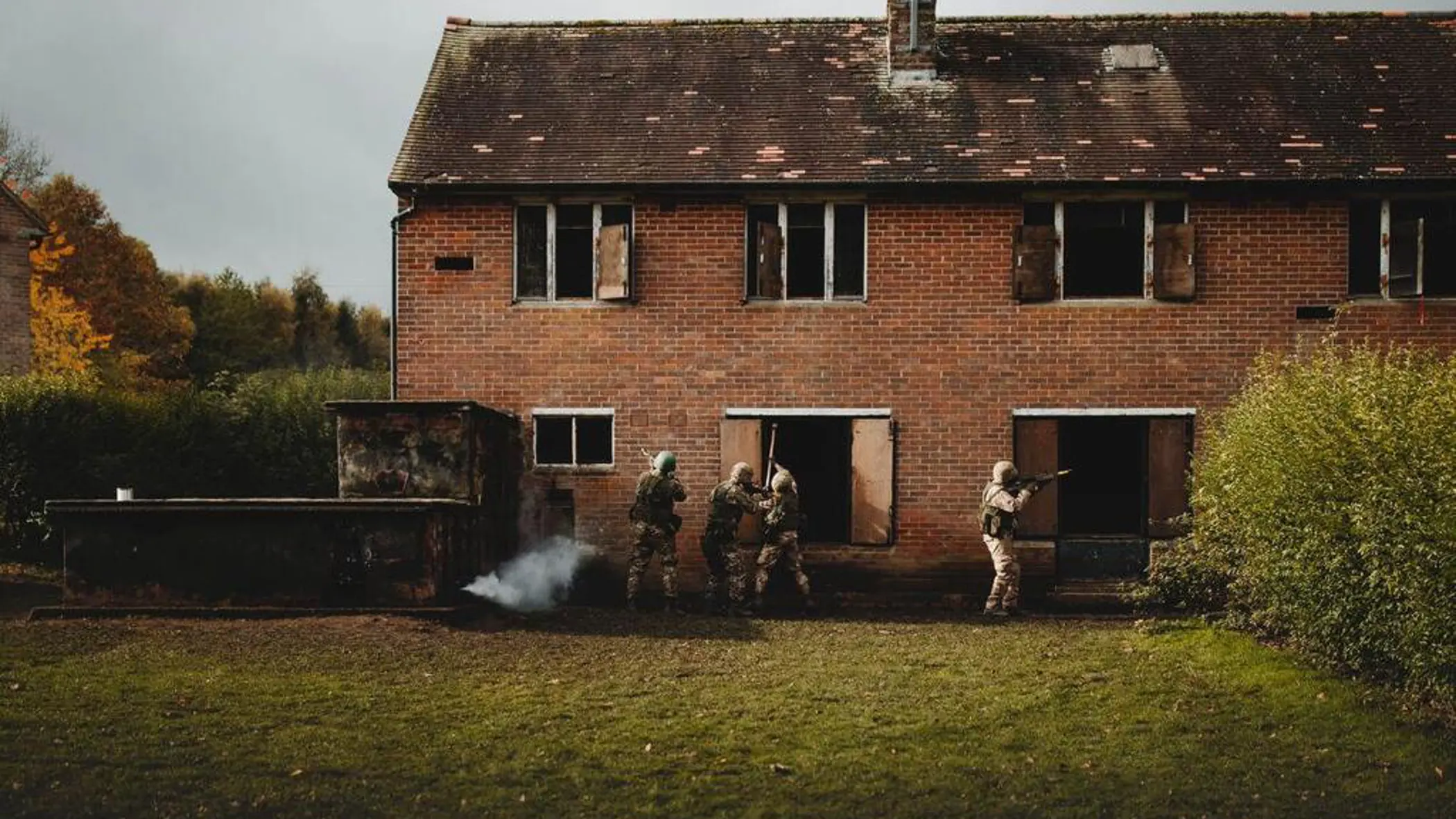 4 soldaten in camouflage benaderen een verlaten bakstenen gebouw, rook komt uit een raam.
