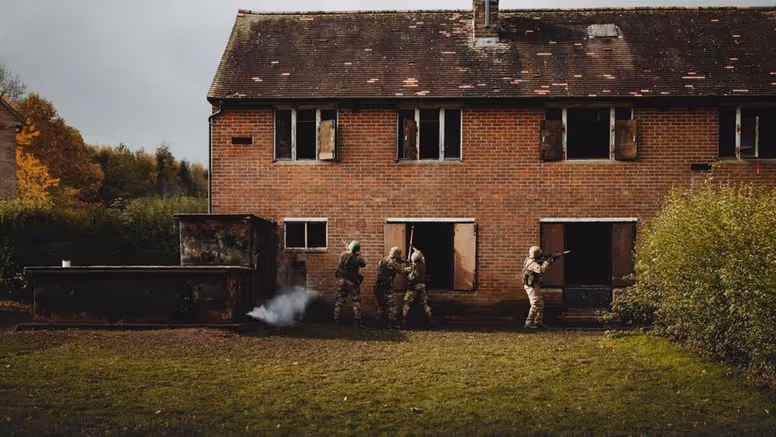 4 soldaten in camouflage benaderen een verlaten bakstenen gebouw, rook komt uit een raam.
