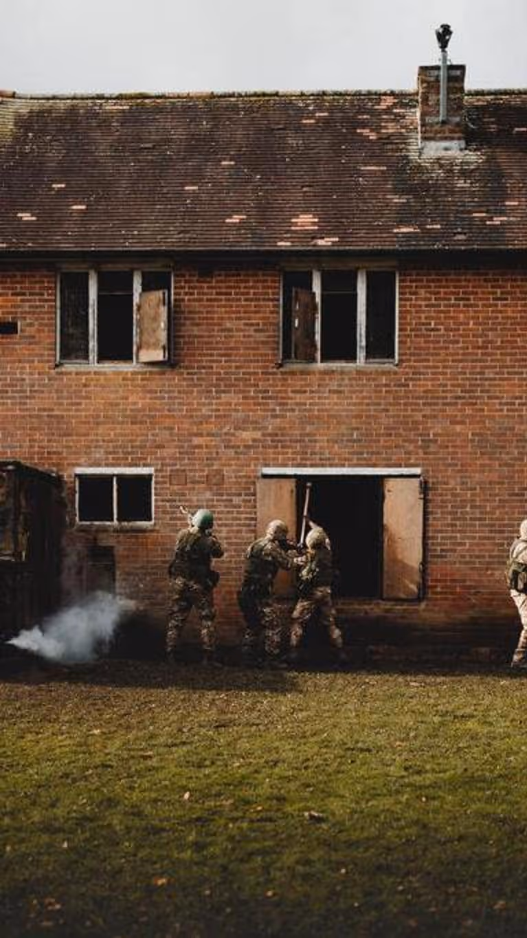 4 soldaten in camouflage benaderen een verlaten bakstenen gebouw, rook komt uit een raam.