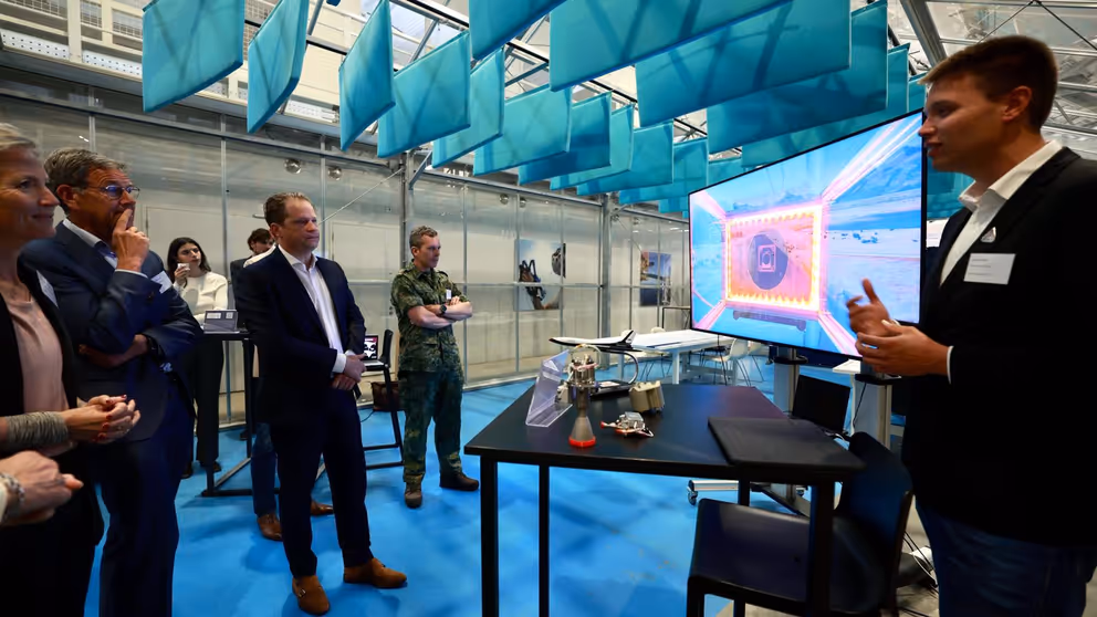 7 mensen in bij een tafeltje en groot scherm. Een man legt iets uit. 