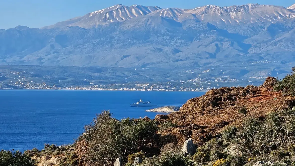 Zr.Ms. De Ruyter ligt in een baai, op de achtergrond zijn bergen zichtbaar.