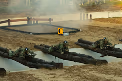 Een greppel met boomstammen die eroverheen liggen waar militairen overheen kruipen en rook oop  de achtergrond.