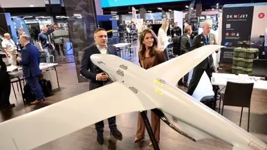 Yeşilgöz op een beursvloer samen met een man in gekleed pak bij een drone. Boven hen een lichtbord met de tekst ‘Drone Summit 2026’.
