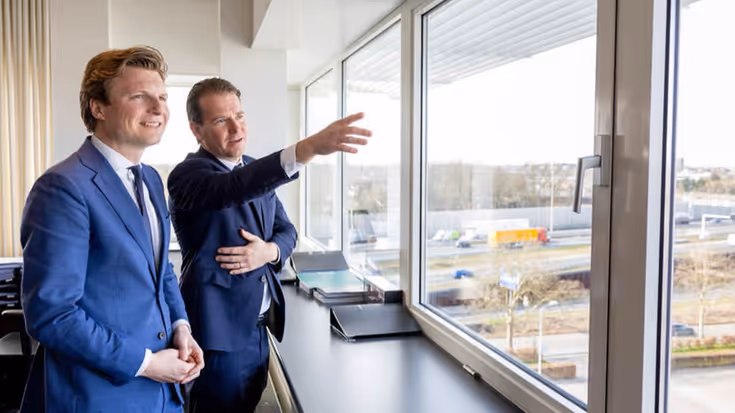 Twee mannen in pak kijken uit het raam over fabrieksterrein. Een van hen wijst iets aan.