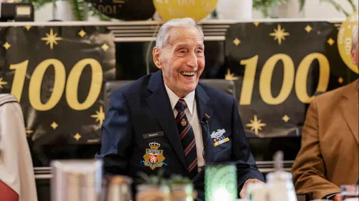 Ton Boelens, zittend aan een tafel, met de cijfers 100 aan beide zijden op de muur erachter.