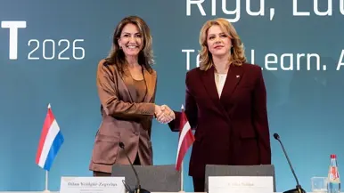 Yeşilgöz en Siliņa schudden elkaar de hand, staand achter een bureau met daarop een kleine Nederlandse en Letse vlag.