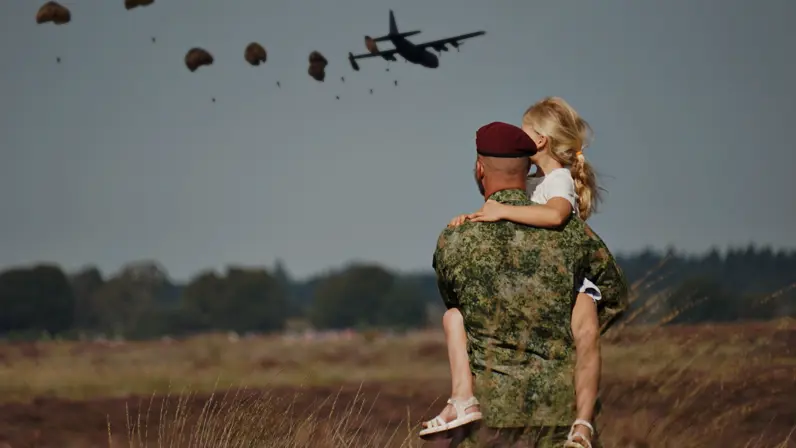 Een militair met een rode baret houdt een blond meisje in zijn armen en kijkt naar parachutisten die net uit een vliegtuig zijn gesprongen.