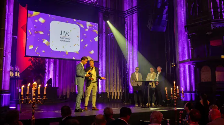 Twee mannen in pak op een podium, van wie een een microfoon vasthoudt. Ze staan in een statige, paars verlichte zaal.