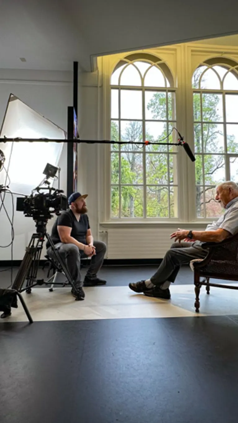 Mijnheer Boes zit op een stoel onder een microfoon. Tegenover hem zit regisseur Joas Burggraaf, die hem interviewt. 
