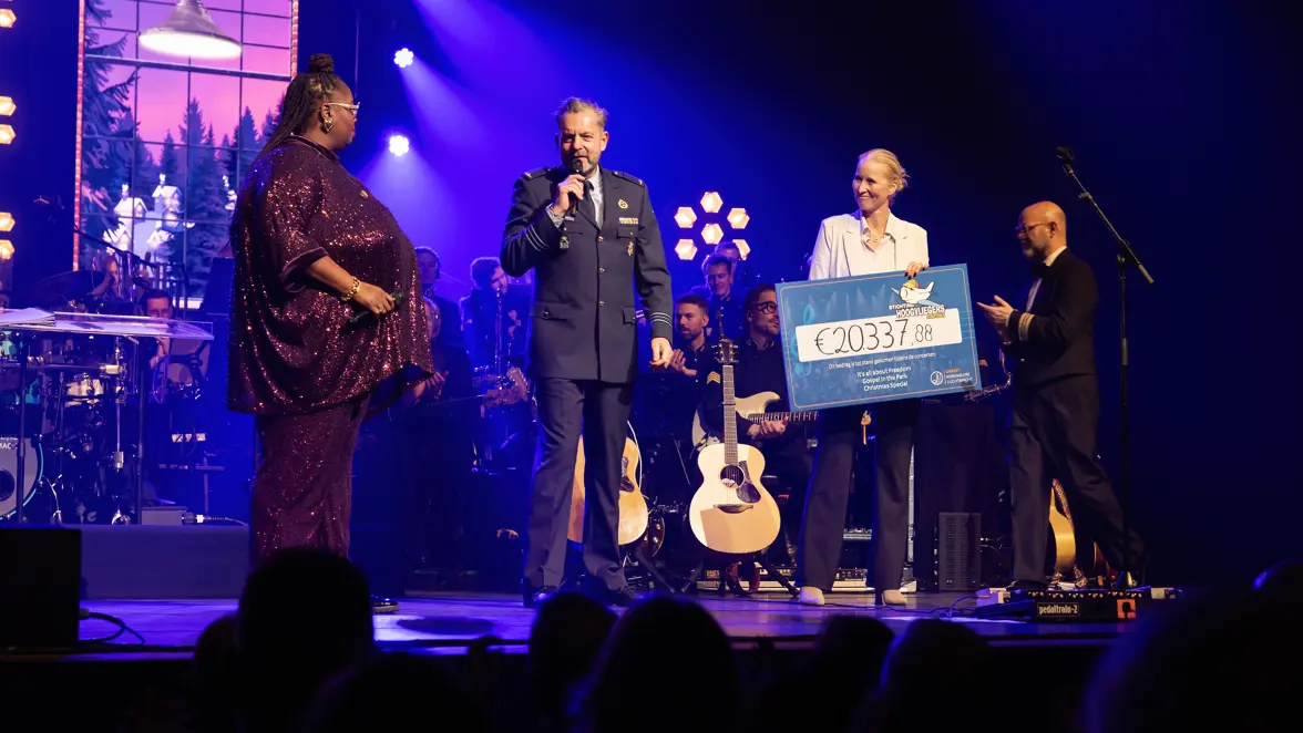 Op het podium staan zangeres Shirma Rouse, majoor Toine Jongbloets pratend door een microfoon en commodore Ellen Meeuwsen-Scholten met in haar handen een grote cheque. Op de achtergrond zit het orkest.