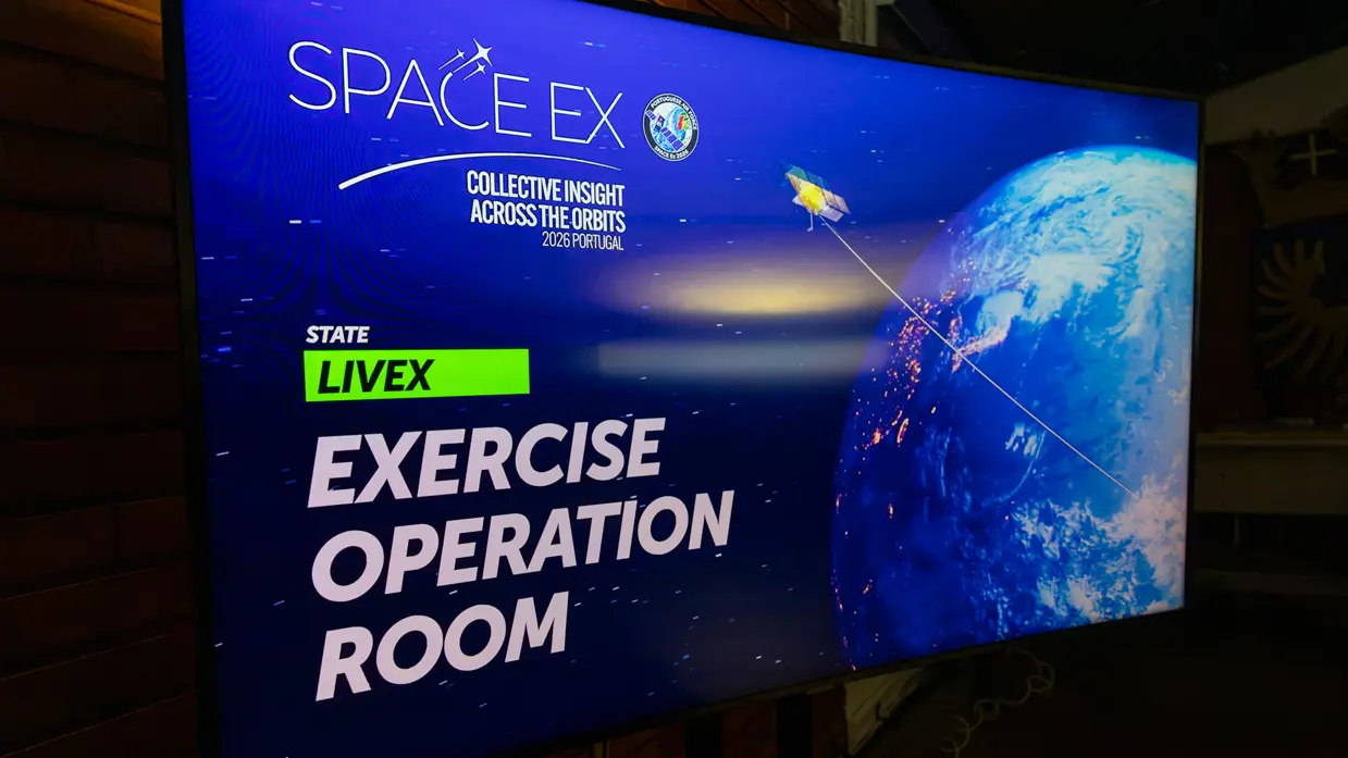 Digitaal scherm met daarop de wereldbol gezien vanuit de ruimte met daarboven een satelliet. Over de afbeelding staat de tekst: ‘SpaceEx; Collective insight across the orbits; Portugal 2026. State LiveEx; Exercise Operation Room’.