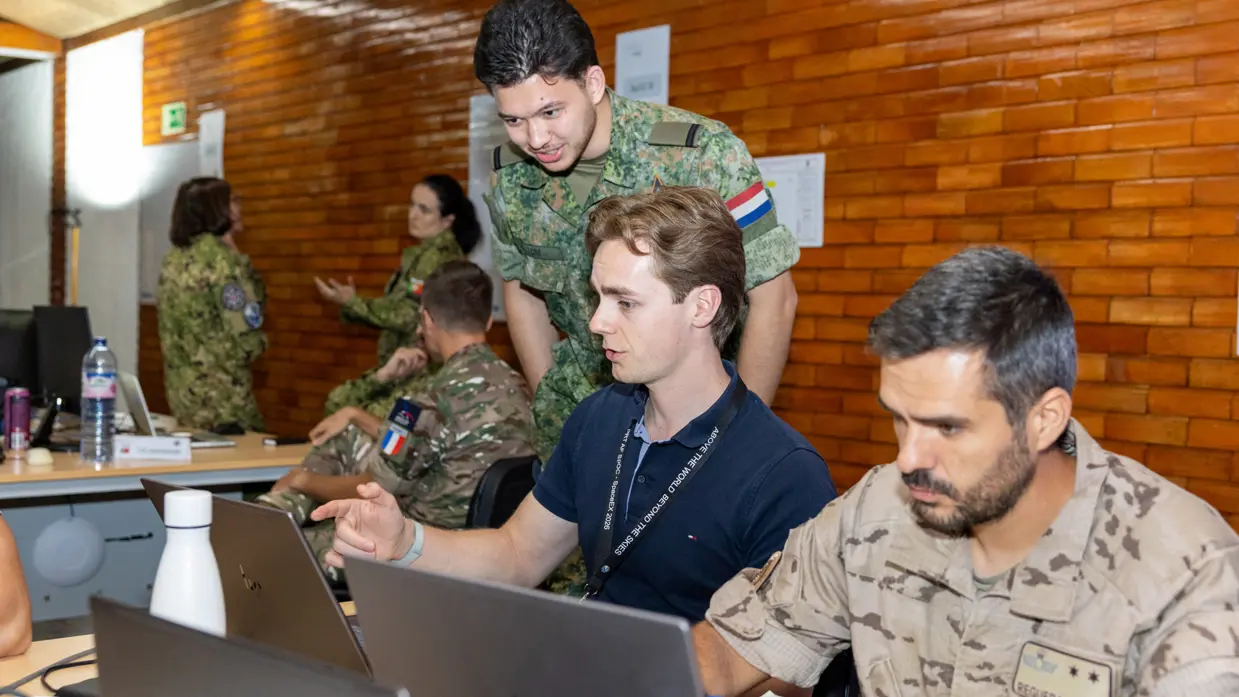 Twee militairen en een burger aan het werk op een paar laptops, een van de militairen is Nederlands. Op de achtergrond nog vier militairen aan het werk.