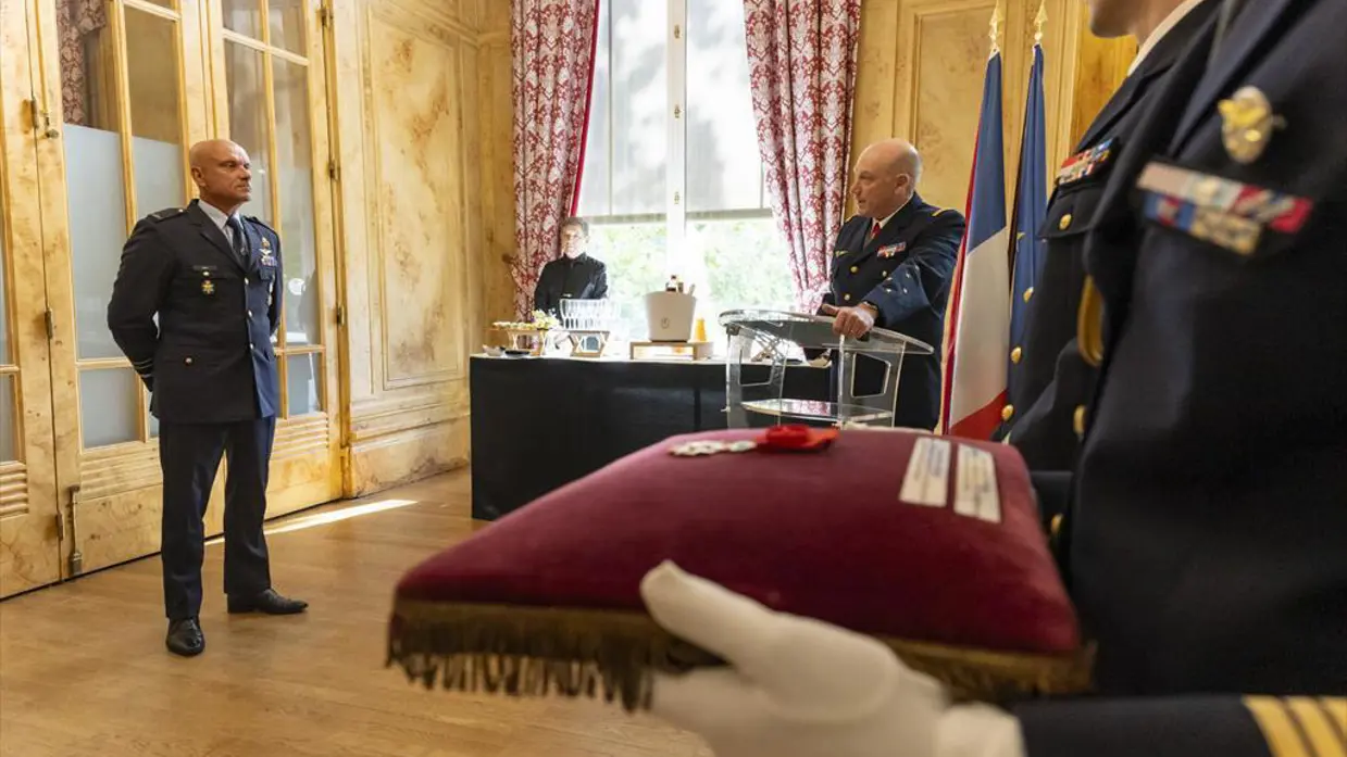  Steur staat ‘op de plaats rust’ in een chique kamer met hoog plafond. Een militair spreekt hem toe vanachter een spreekgestoelte. Een ander militair houd een bordeaux rood kussen vast met daarop een medaille.