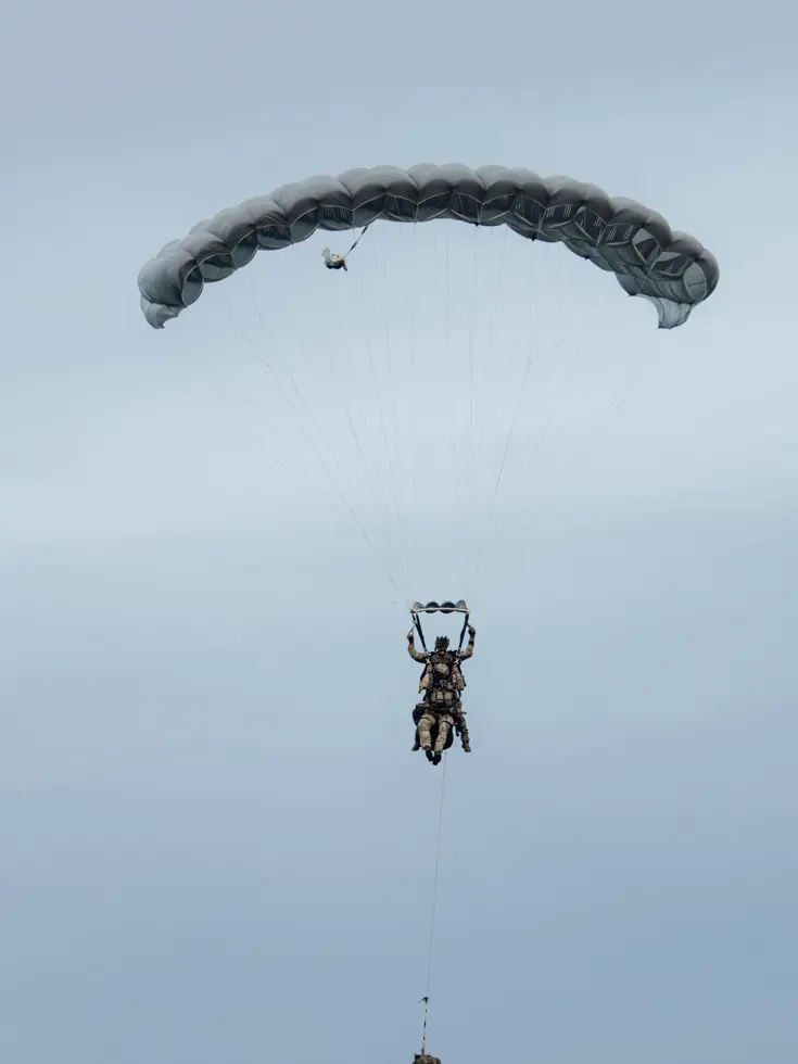 Een parachutist daalt neer uit de lucht met volledig geopende parachute. Aan hem vast zit nog een andere militair.