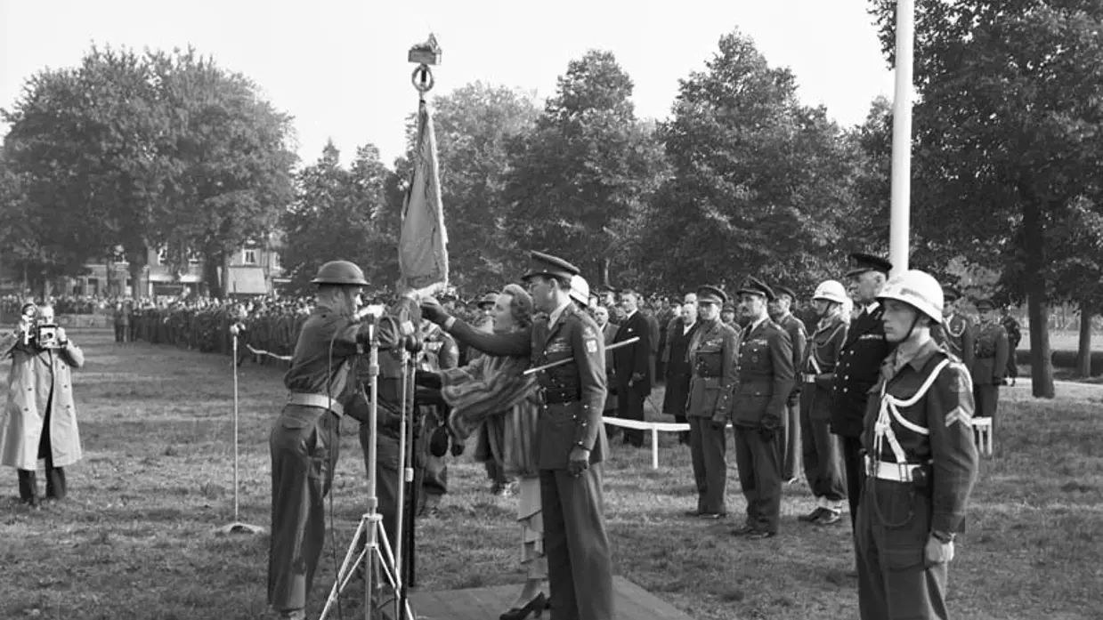 Militairen staan opgesteld. 2 militairen dragen vaandel aan elkaar over.