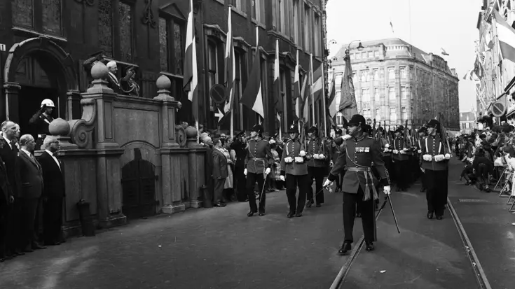 Zwart-witfoto militairen in ceremonieel tenue marcheren over straat.